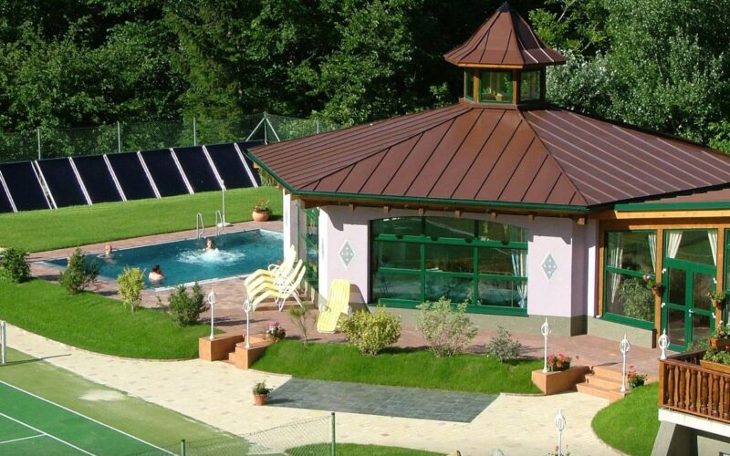 Ein kleiner Swimmingpool mit Liegestühlen befindet sich neben einem Pavillon mit großen Fenstern und einem braunen Dach, umgeben von Grünanlagen und einem Tennisplatz.