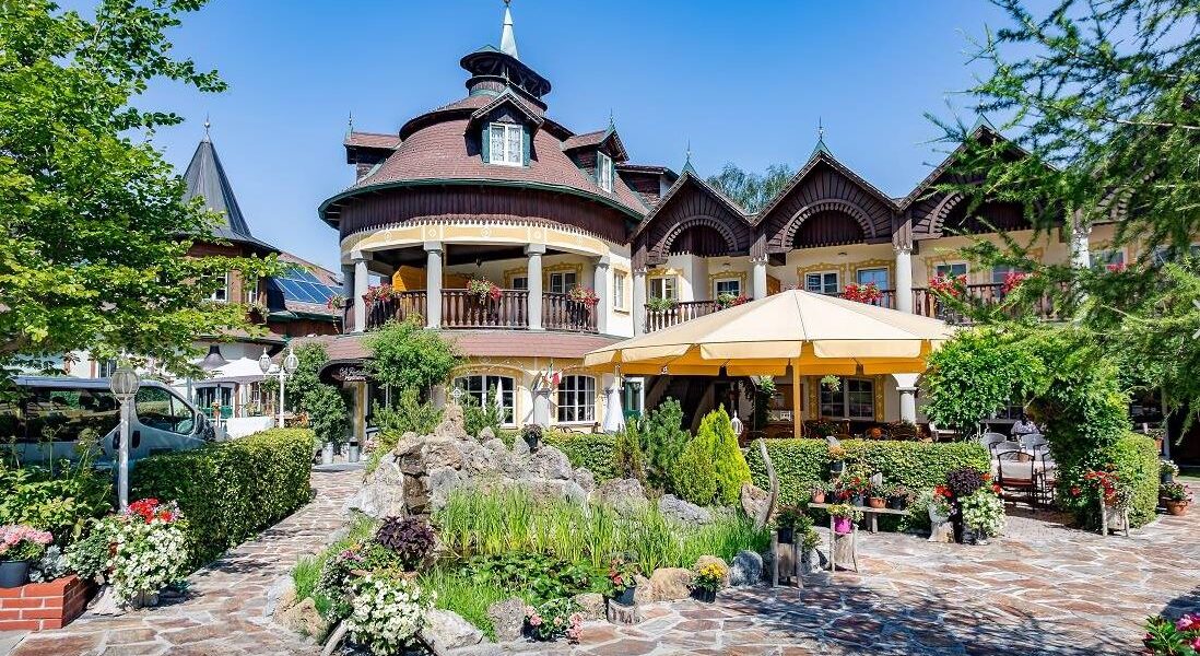 Ein großes, verziertes Gebäude mit Holzakzenten und Balkonen, umgeben von üppigen Gärten, einer Steinterrasse, einem Teich und Sitzgelegenheiten im Freien unter einem gelben Sonnenschirm.