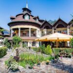 Ein großes, verziertes Gebäude mit Holzakzenten und Balkonen, umgeben von üppigen Gärten, einer Steinterrasse, einem Teich und Sitzgelegenheiten im Freien unter einem gelben Sonnenschirm.