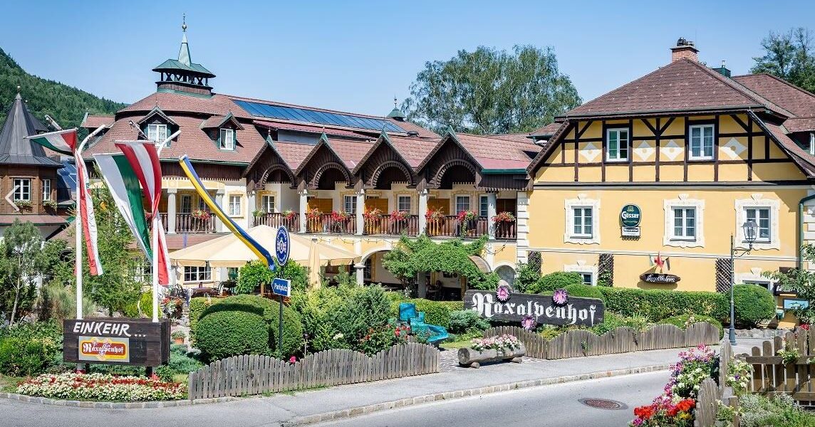 Ein traditionelles gelbes Alpenhotel mit Balkonen und Fahnen steht an einer Straße, umgeben von Grün und dekorativen Schildern, unter einem klaren Himmel.