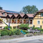 Ein traditionelles gelbes Alpenhotel mit Balkonen und Fahnen steht an einer Straße, umgeben von Grün und dekorativen Schildern, unter einem klaren Himmel.