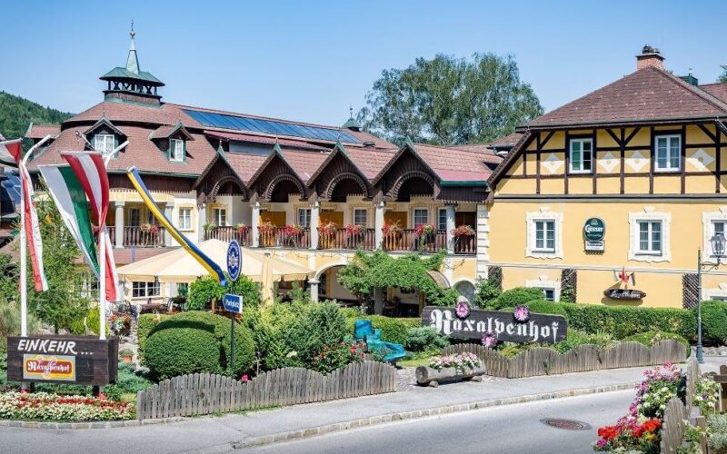 Ein traditionelles gelbes Alpenhotel mit Balkonen und Fahnen steht an einer Straße, umgeben von Grün und dekorativen Schildern, unter einem klaren Himmel.