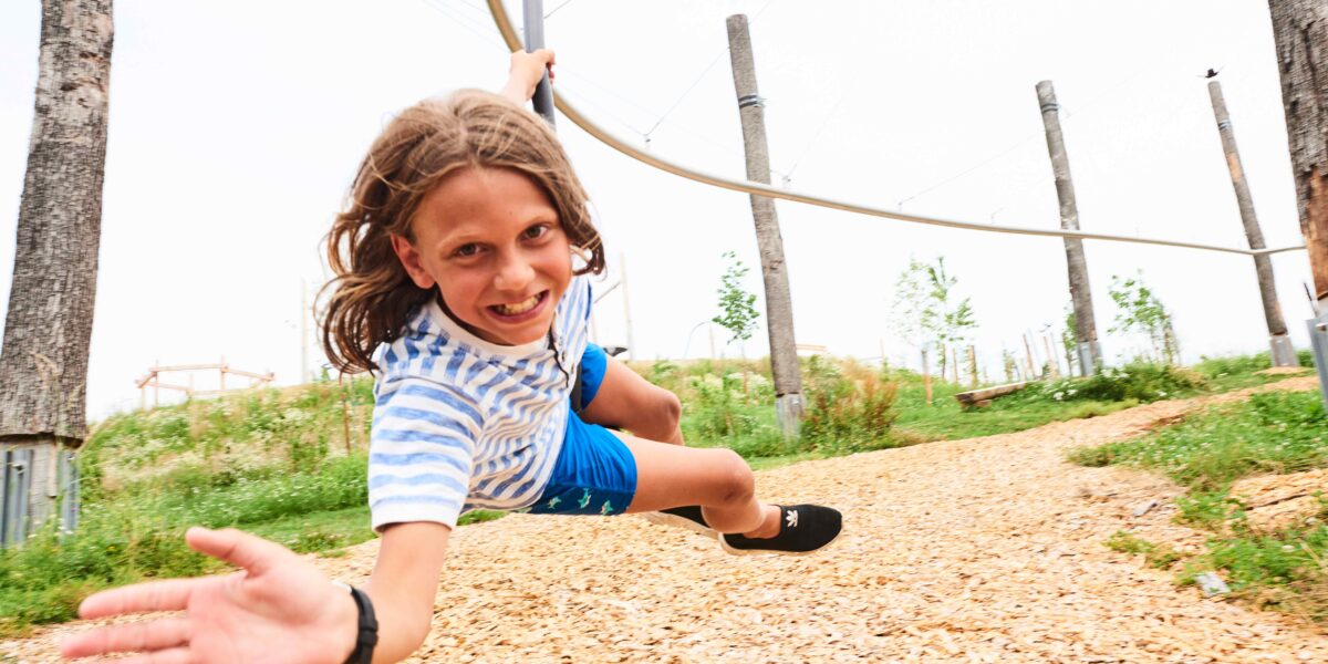Ein Kind in einem gestreiften Hemd und blauen Shorts fährt auf einer Seilrutsche im Freien, lächelt und streckt eine Hand nach vorne, während Holzspäne auf dem Boden liegen.