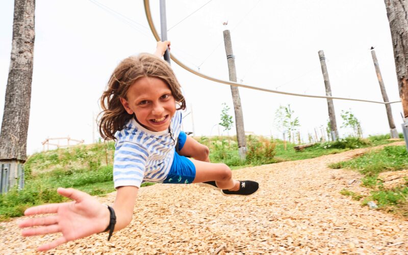 Ein Kind in einem gestreiften Hemd und blauen Shorts fährt auf einer Seilrutsche im Freien, lächelt und streckt eine Hand nach vorne, während Holzspäne auf dem Boden liegen.
