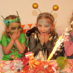 Fünf kleine Kinder in Kostümen sitzen überrascht und aufgeregt an einem Tisch und betrachten eine Torte mit einer Wunderkerze auf einer Geburtstagsparty.