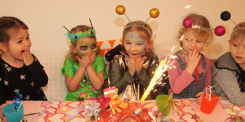 Fünf kleine Kinder in Kostümen sitzen überrascht und aufgeregt an einem Tisch und betrachten eine Torte mit einer Wunderkerze auf einer Geburtstagsparty.