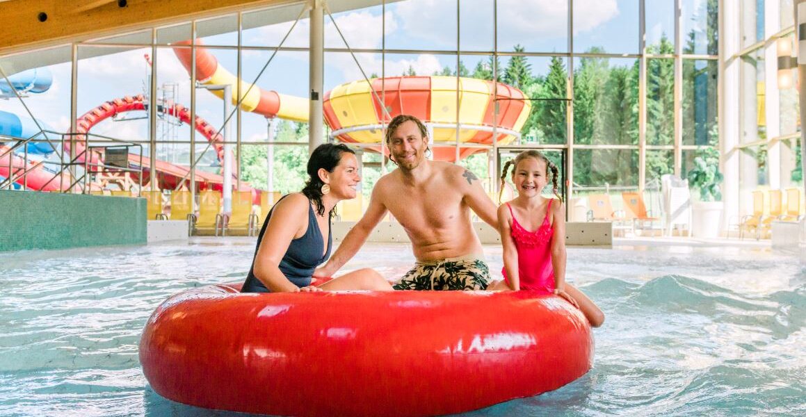 Ein Mann und zwei Kinder sitzen auf einem großen roten aufblasbaren Ring in einem Indoor-Wasserpark, mit Wasserrutschen und großen Fenstern im Hintergrund.