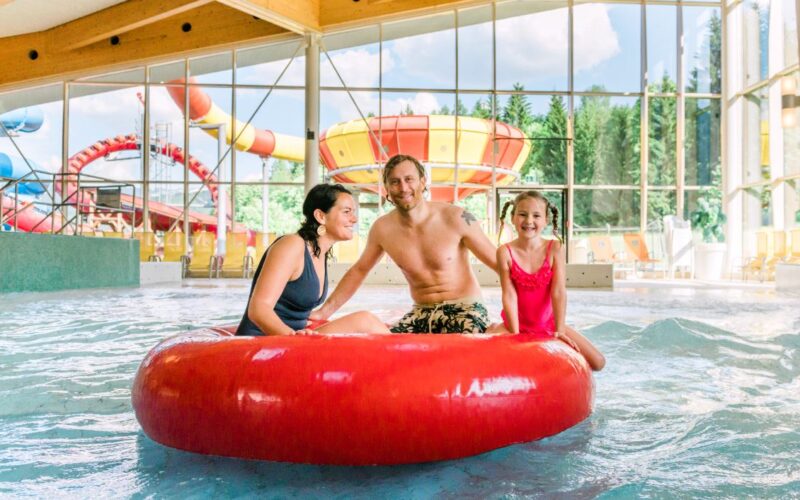 Ein Mann und zwei Kinder sitzen auf einem großen roten aufblasbaren Ring in einem Indoor-Wasserpark, mit Wasserrutschen und großen Fenstern im Hintergrund.
