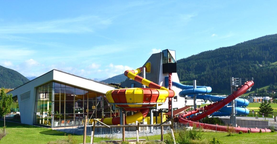 Ein modernes Wasserparkgebäude mit großen Glasfenstern und mehreren farbenfrohen Wasserrutschen im Freien vor einer Kulisse aus grünen Hügeln und blauem Himmel.