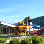 Ein modernes Wasserparkgebäude mit großen Glasfenstern und mehreren farbenfrohen Wasserrutschen im Freien vor einer Kulisse aus grünen Hügeln und blauem Himmel.