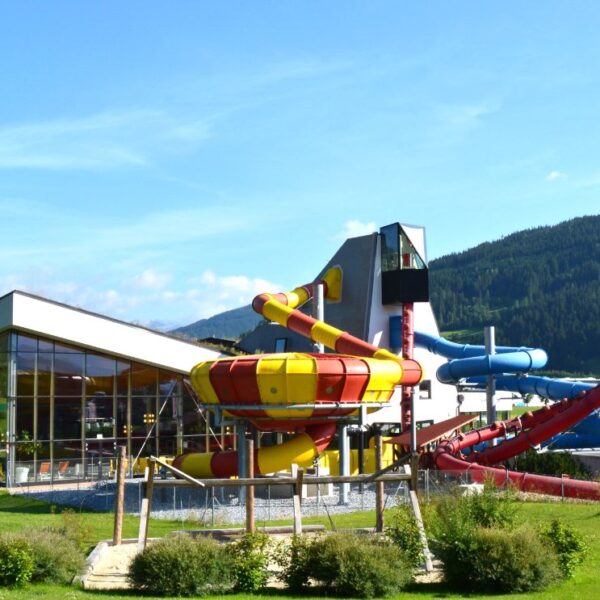 Ein modernes Wasserparkgebäude mit großen Glasfenstern und mehreren farbenfrohen Wasserrutschen im Freien vor einer Kulisse aus grünen Hügeln und blauem Himmel.