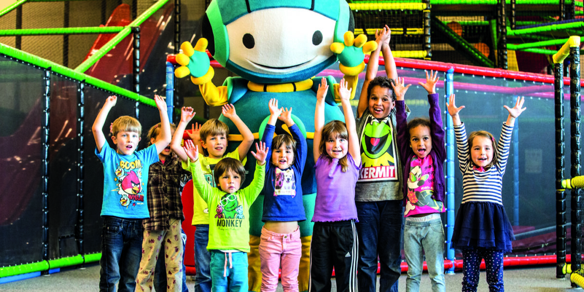 Neun Kinder stehen mit erhobenen Armen vor einem großen, bunten Maskottchen in einem Indoor-Spielbereich mit Klettergerüsten und Netzen.