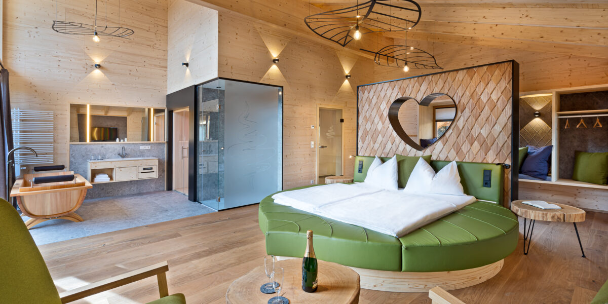 Moderne Hotelsuite mit holzgetäfelten Wänden, rundem Bett mit herzförmigem Kopfteil, offenem Badezimmer, grünen Stühlen und einem kleinen Tisch mit Champagner und Gläsern.
