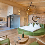 Moderne Hotelsuite mit holzgetäfelten Wänden, rundem Bett mit herzförmigem Kopfteil, offenem Badezimmer, grünen Stühlen und einem kleinen Tisch mit Champagner und Gläsern.