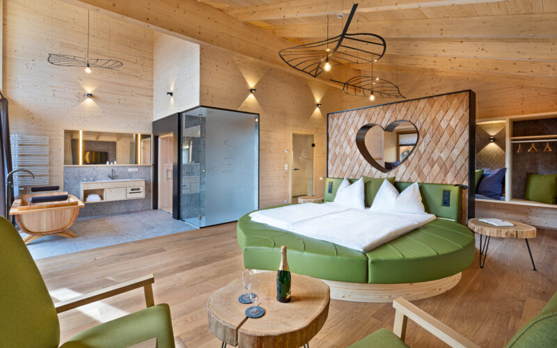 Moderne Hotelsuite mit holzgetäfelten Wänden, rundem Bett mit herzförmigem Kopfteil, offenem Badezimmer, grünen Stühlen und einem kleinen Tisch mit Champagner und Gläsern.