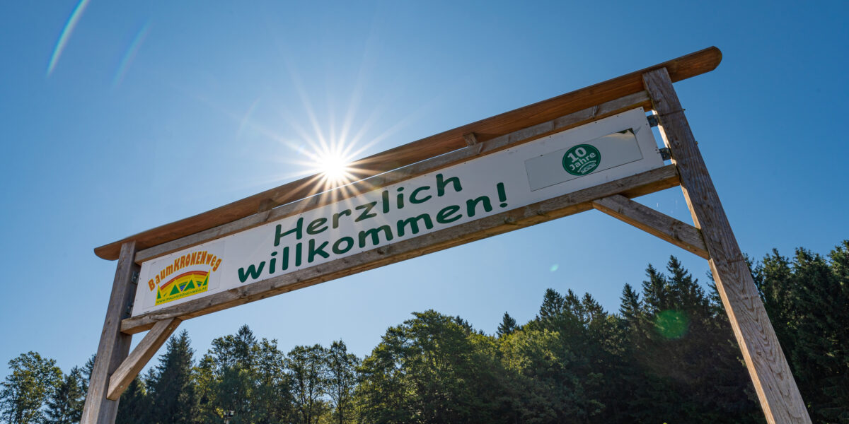Ein großes Holzschild mit der Aufschrift „Herzlich willkommen!“ steht vor einem klaren blauen Himmel mit Sonnenlicht und Bäumen im Hintergrund.