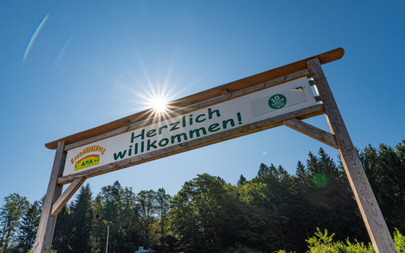 Ein großes Holzschild mit der Aufschrift „Herzlich willkommen!“ steht vor einem klaren blauen Himmel mit Sonnenlicht und Bäumen im Hintergrund.