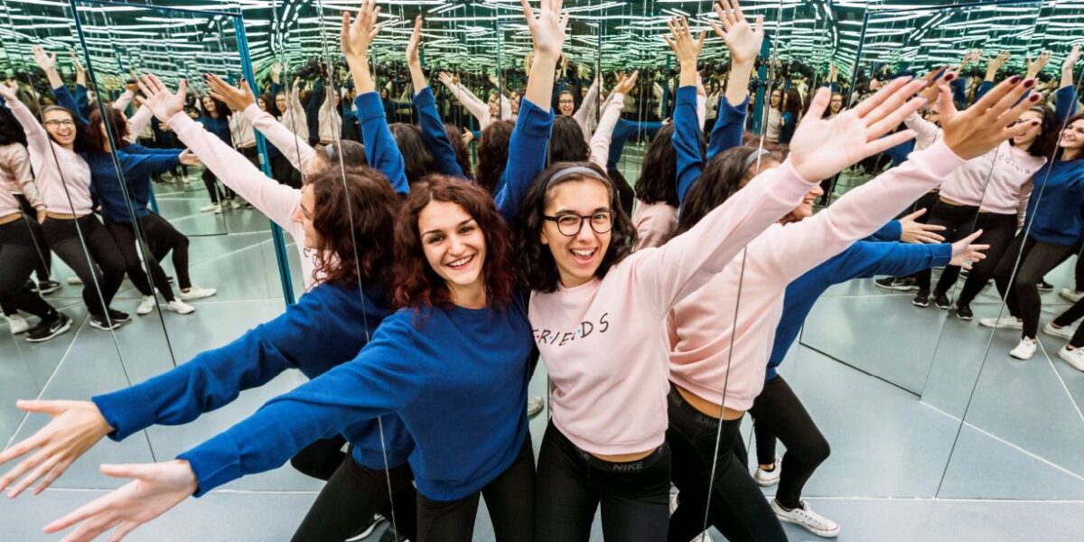Zwei Personen stehen mit ausgestreckten Armen in einem verspiegelten Raum und spiegeln sich mehrfach an den Wänden und der Decke.