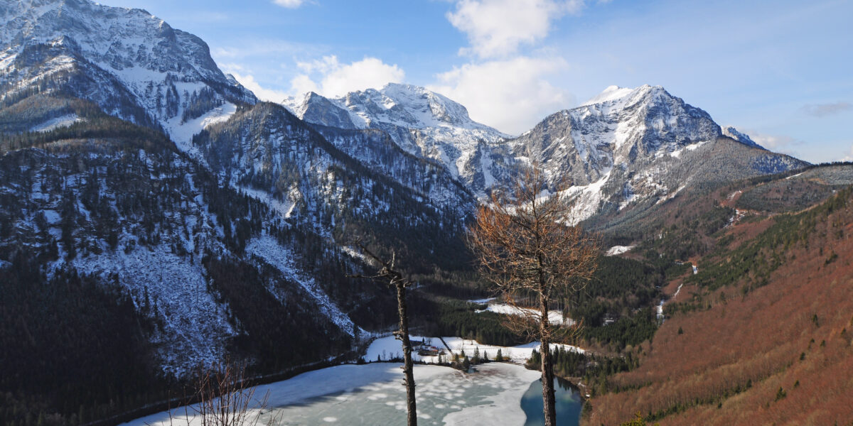 Ein teilweise zugefrorener See, umgeben von schneebedeckten Bergen und bewaldeten Hängen unter einem blauen Himmel mit vereinzelten Wolken.