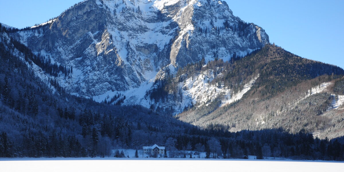 Schneebedeckter Berg mit bewaldetem Fuß und einem Haus im Vordergrund unter einem klaren blauen Himmel.