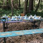 Ein Picknicktisch in einem Wald ist für eine Kinderparty mit bunten Hüten, Tellern, Tassen, Snacks und Getränken auf einer abgenutzten Holzbank und einem Tisch gedeckt.