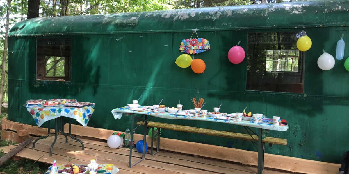 Dekorierte Party im Freien mit Tischen, Essen und Luftballons vor einem grünen, von Bäumen umgebenen Zugwaggon.