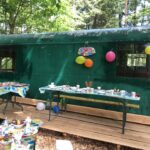 Dekorierte Party im Freien mit Tischen, Essen und Luftballons vor einem grünen, von Bäumen umgebenen Zugwaggon.