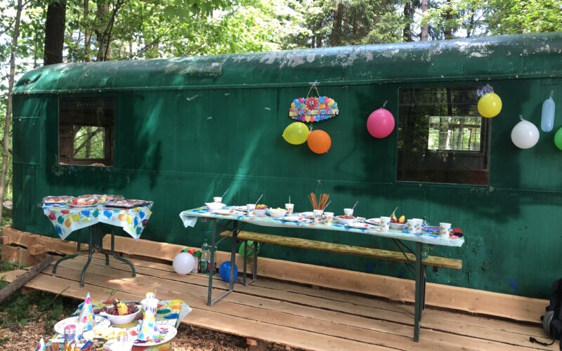 Dekorierte Party im Freien mit Tischen, Essen und Luftballons vor einem grünen, von Bäumen umgebenen Zugwaggon.