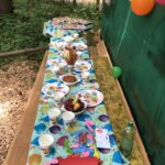 Ein langer Picknicktisch in einem Waldgebiet ist mit bunten Partytellern, Bechern, Snacks, Luftballons und Utensilien gedeckt, bereit für eine Kinderfeier im Freien.