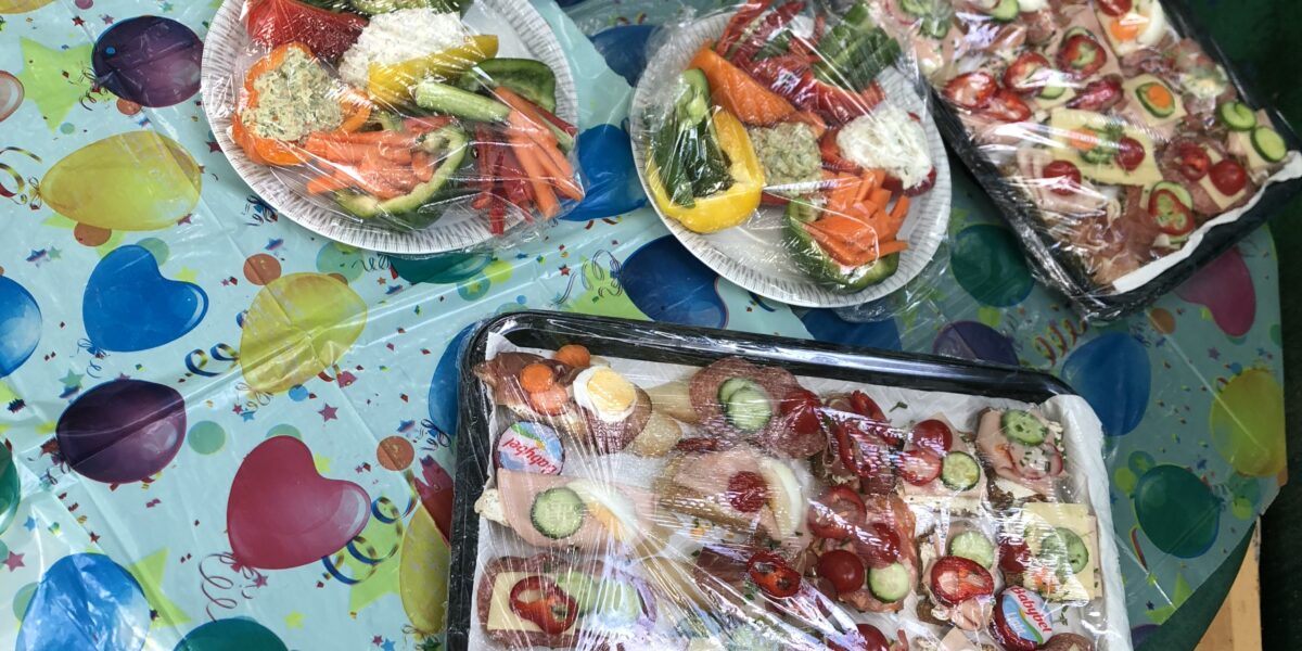 Auf einem runden Tisch, der mit einem bunten, mit Luftballons bedruckten Tuch bedeckt ist, stehen verschiedene Tabletts mit Aufschnitt, Gemüse, Käse und in Plastik eingewickelten Sandwiches.