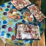 Auf einem runden Tisch, der mit einem bunten, mit Luftballons bedruckten Tuch bedeckt ist, stehen verschiedene Tabletts mit Aufschnitt, Gemüse, Käse und in Plastik eingewickelten Sandwiches.