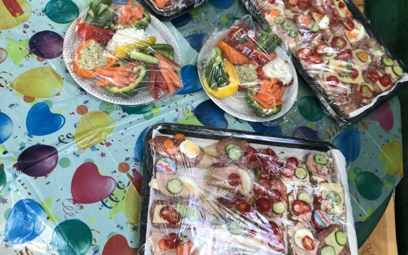 Auf einem runden Tisch, der mit einem bunten, mit Luftballons bedruckten Tuch bedeckt ist, stehen verschiedene Tabletts mit Aufschnitt, Gemüse, Käse und in Plastik eingewickelten Sandwiches.