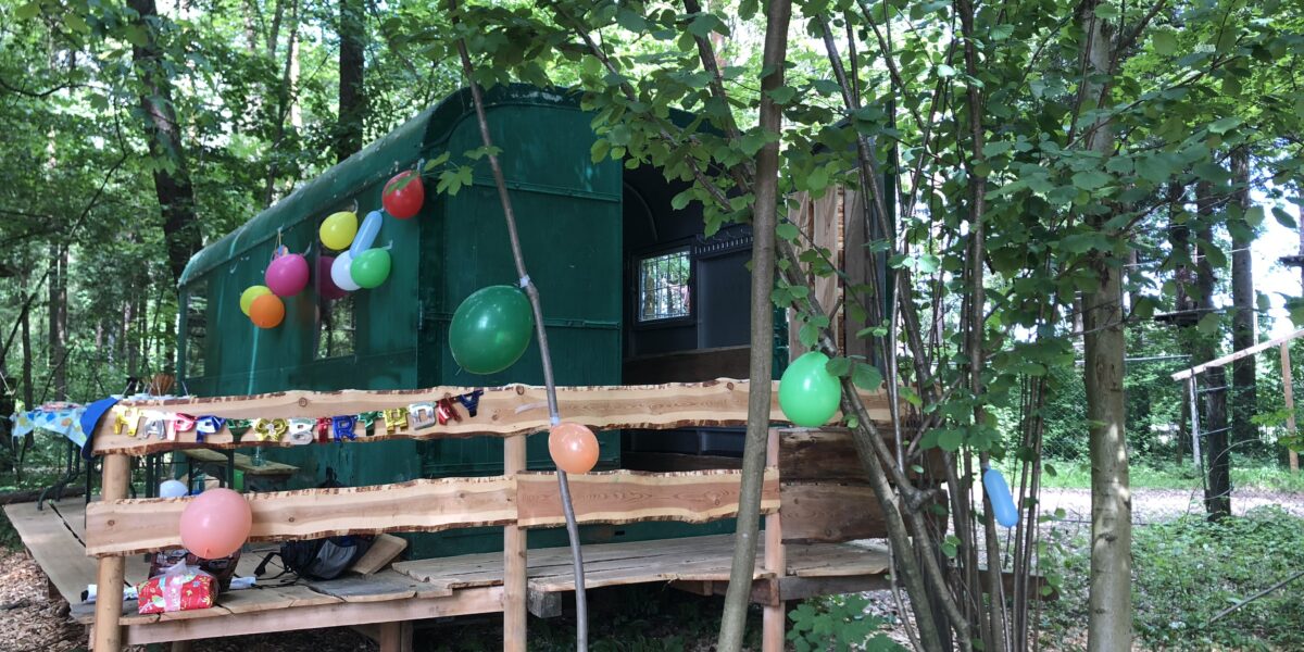 Ein grünes, zeltartiges Gebäude im Wald ist mit bunten Luftballons und einem Geburtstagsbanner geschmückt. Eine hölzerne Rampe mit Geländer führt zum Eingang hinauf.