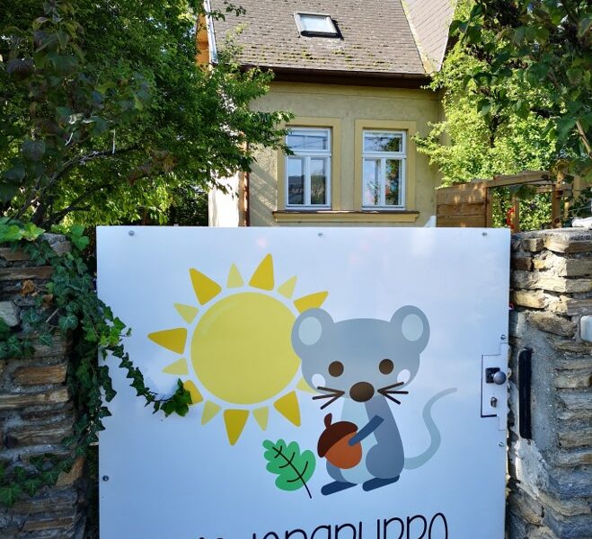Ein Schild mit einer Cartoon-Maus, einer Sonne und der Aufschrift "Kindergruppe Sonnenmaus" ist an einem Tor vor einem von Bäumen umgebenen Haus angebracht.