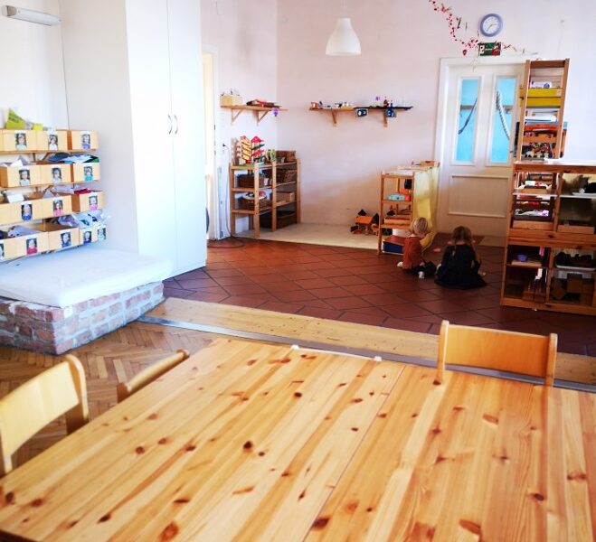 Ein Klassenzimmer mit Holztischen und -stühlen im Vordergrund, Regalen mit Spielzeug und zwei Kindern, die in der Nähe der Rückwand auf dem Boden spielen.