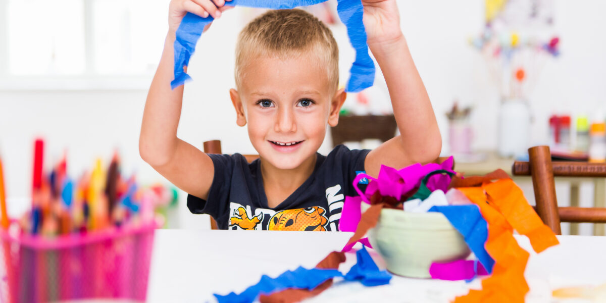 Ein lächelnder Junge hält einen Streifen blaues Krepppapier an einem Tisch mit buntem Bastelmaterial und Papier in einem hellen Raum hoch.