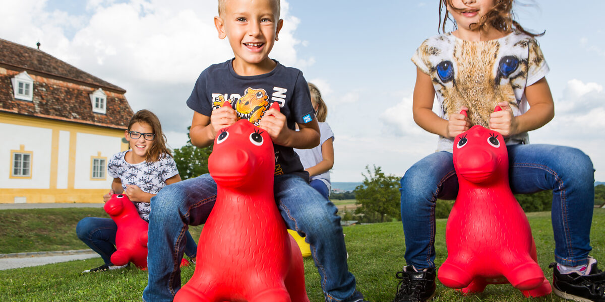 Vier Kinder spielen im Freien, sitzen auf roten aufblasbaren Tierspielzeugen auf einer Wiese in der Nähe eines Gebäudes mit einem Ziegeldach.