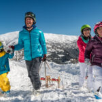 Eine vierköpfige Familie in Winterkleidung geht in einer verschneiten Berglandschaft bergauf und zieht einen Schlitten unter einem strahlend blauen Himmel.