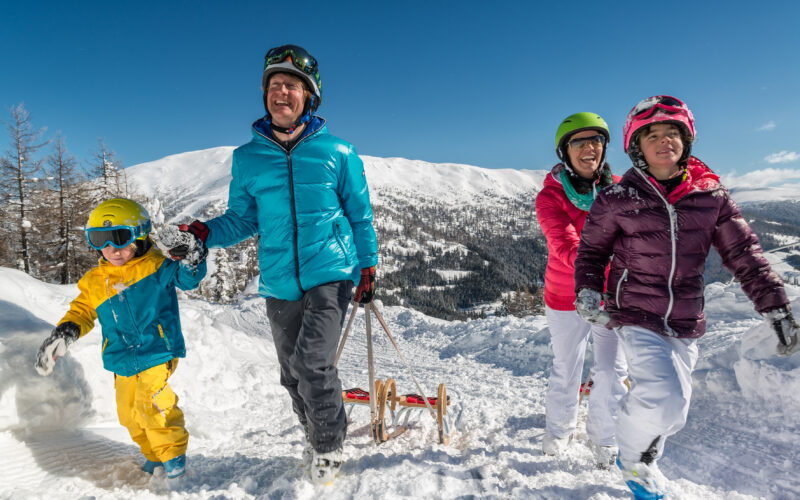 Eine vierköpfige Familie in Winterkleidung geht in einer verschneiten Berglandschaft bergauf und zieht einen Schlitten unter einem strahlend blauen Himmel.
