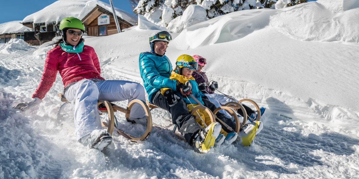 Vier Personen, zwei Erwachsene und zwei Kinder, sitzen auf Holzschlitten im Schnee, tragen Helme und Winterkleidung, im Hintergrund verschneite Bäume und eine Hütte.