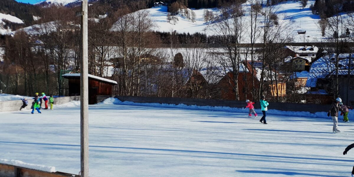 Menschen beim Schlittschuhlaufen auf einer Freiluft-Eisbahn, umgeben von Schnee, Bäumen und Bergen unter blauem Himmel.