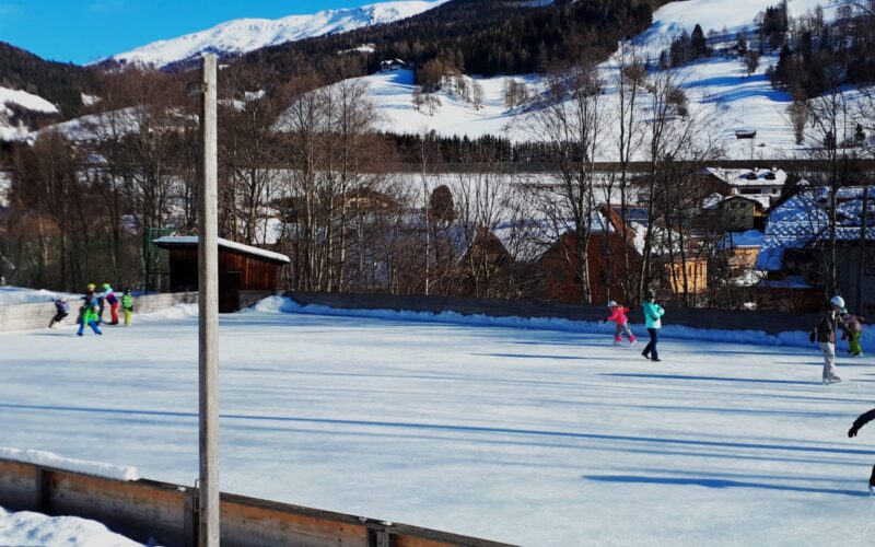 Menschen beim Schlittschuhlaufen auf einer Freiluft-Eisbahn, umgeben von Schnee, Bäumen und Bergen unter blauem Himmel.