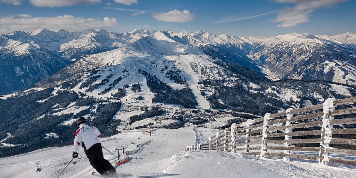 Ein Skifahrer in einer weißen Jacke fährt auf einer verschneiten Bergpiste bergab, mit einem Holzzaun auf der rechten Seite und schneebedeckten Gipfeln im Hintergrund unter einem teilweise bewölkten Himmel.