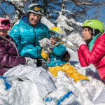 Vier Menschen in bunten Winterklamotten sitzen im Schnee, lächeln und spielen zusammen mit schneebedeckten Bäumen im Hintergrund.
