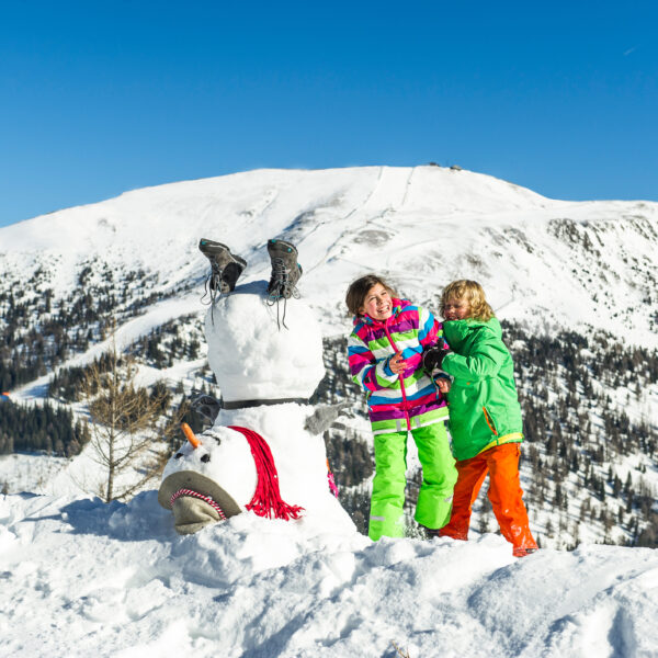 Zwei Kinder in farbenfroher Winterkleidung lachen neben einem umgedrehten Schneemann auf einem verschneiten Berg unter einem klaren blauen Himmel.