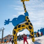 Kinder beim Skifahren unter einer großen gelben und blauen aufblasbaren Giraffenstruktur auf einer verschneiten Piste mit Skiliften und Gebäuden im Hintergrund.
