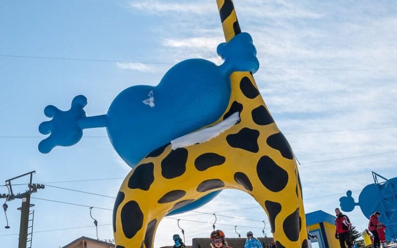 Kinder beim Skifahren unter einer großen gelben und blauen aufblasbaren Giraffenstruktur auf einer verschneiten Piste mit Skiliften und Gebäuden im Hintergrund.