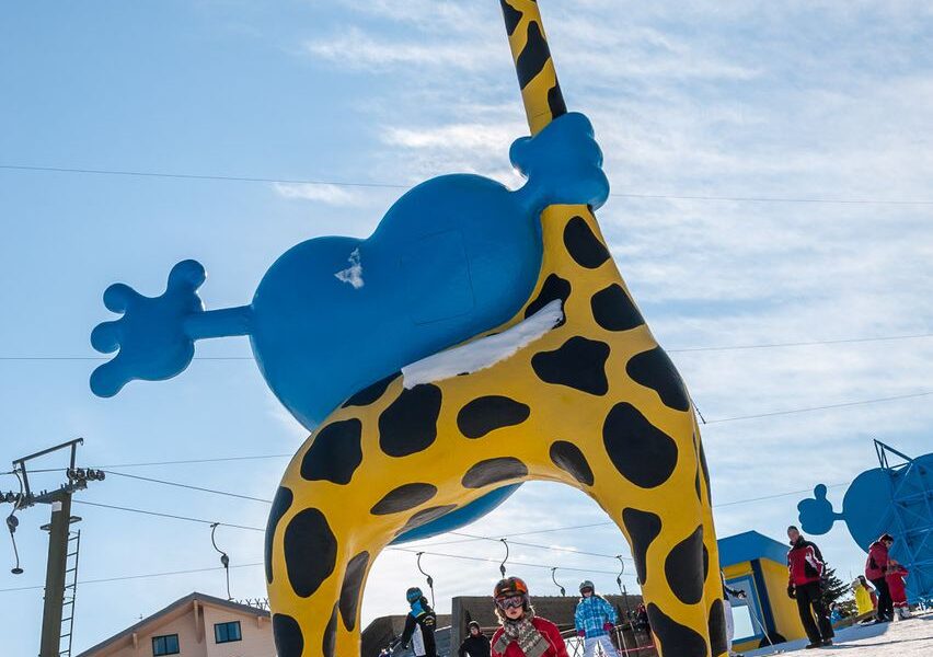 Kinder beim Skifahren unter einer großen gelben und blauen aufblasbaren Giraffenstruktur auf einer verschneiten Piste mit Skiliften und Gebäuden im Hintergrund.