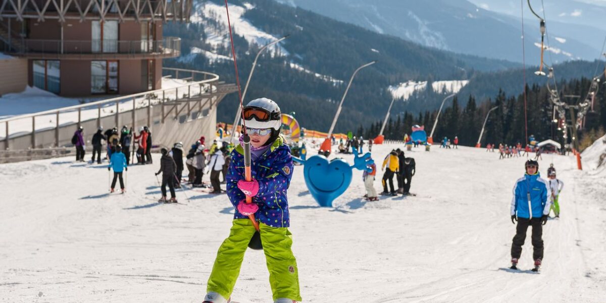 Ein Kind in Skiausrüstung hält ein Schleppseil auf einer verschneiten Piste, im Hintergrund sind andere Skifahrer und ein Gebäude zu sehen, in der Ferne die Berge.