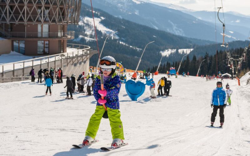 Ein Kind in Skiausrüstung hält ein Schleppseil auf einer verschneiten Piste, im Hintergrund sind andere Skifahrer und ein Gebäude zu sehen, in der Ferne die Berge.